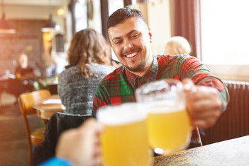 First person portrait of a young cheerful smiling man in casual wear toasting with beer/ale while sitting in pub