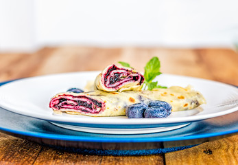 Lateral view of crepe with blueberry jam filling cut in half and decorated with mint leaves and frozen blueberries on white and blue plates.
