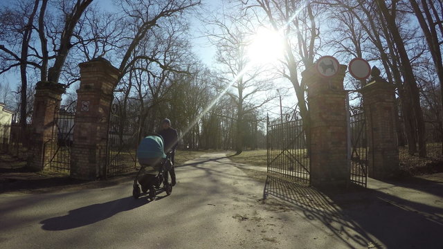 father man with blue stroller walk through retro park gate entrance in sunny spring day. Wide angle shot. 4K UHD.