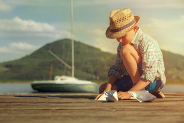 Fototapeta premium Little boy make paper boats sitting on the wooden pier
