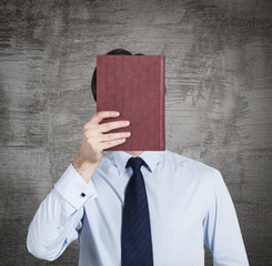 A person holds a red book in front of the head. Concrete background.
