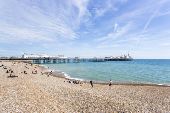 The Shingle Beach At Brighton, East Sussex, UK In Summer And The Palace Pier (Brighton Pier) With Holidaymakers Sunbathing On A Sunny Day Under A Blue Sky And Wispy Clouds