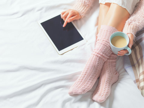 Woman Using Tablet At Cozy Soft Home Atmosphere On The Bed With Cup Of Coffee Or Cocoa. 