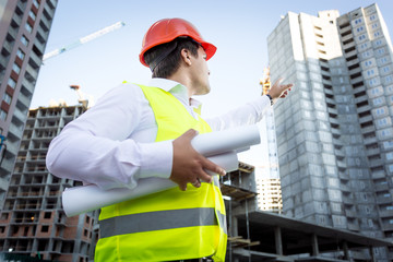 portrait of foreman with blueprints pointing hand at building