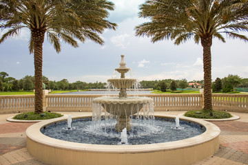 Fototapeta premium Fountain and Lake - Water Splashes from Tropical Fountain that Overlooks Lake Surrounded by Lush Greenery