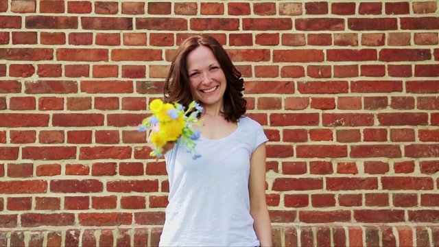 h&uuml;bsche Frau h&auml;lt sch&ouml;ne Blumen in die Kamera, im Hintergrund ist eine Backsteinwand