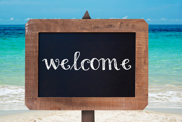 Welcome written on a wooden vintage chalk board, beach background © Delphotostock