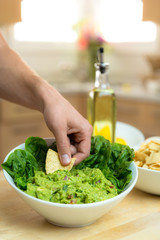 Hand dipping chip into a fresh delicious bowl of handmade guacamole with ingredients nearby