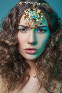 Beautiful fashion model with elegant gold middle eastern headpiece on head looking at camera with serious face and curly hair. on blue background. 
RAW - retouched with special care and attention.
