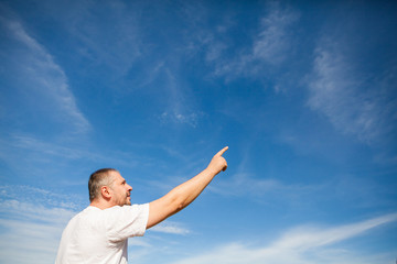 Side portrait low angle view of a man with beard standing and looking ahead against a blue sky with white clouds and pointing with finger