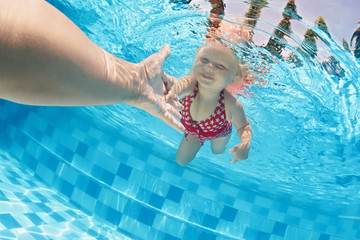 Joyful baby girl diving underwater with fun and holding parents hand for assistance in swimming pool. Healthy active family lifestyle, children water sport activity with mother on summer vacation