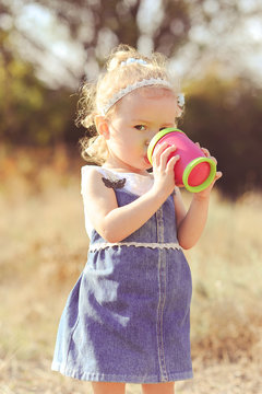 Baby Girl 2-3 Year Old Drinking Water From Plastic Cup Outdoors. Wearing Stylish Denim Dress. Childhood.