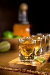 Vertical Close up of Tequila shot glasses grouped together with a bottle in the background and cut limes on a wooden surface