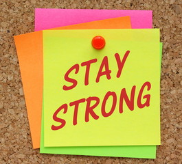 The phrase Stay Strong in red text on a yellow sticky note pinned to a cork notice board