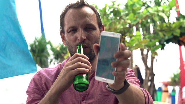 Young man with beer taking selfie photo with cellphone in beach bar, slow motion shot at 240fps
