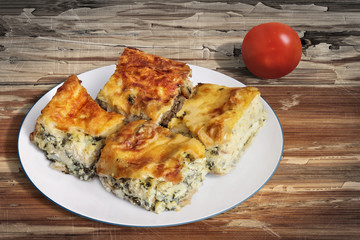 Plate of Freshly baked Serbian Traditional Zeljanica Spinach-cheese Pie slices, with Tomato, set on very old, weathered, cracked, scratched, peeled off, Wooden Table surface.