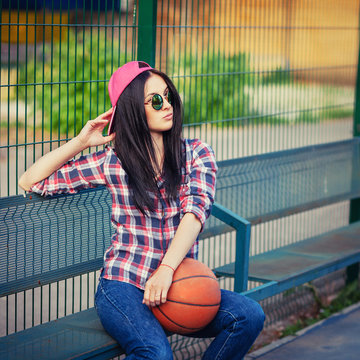 Outdoor lifestyle portrait of pretty young sitting girl, wearing in hipster swag grunge style with basketball in urban background. Retro vintage toned image, film simulation.