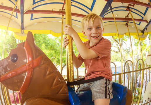 Cute little smiling boy riding on a Carnival Carousel at an amusement park or theme park. Warm afternoon sun in the background