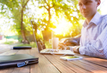 Businessman working outdoor.
Smart casual dressed person working on computer drinking coffee mug sitting at rough natural wooden desk outdoor with green tree and sun on background