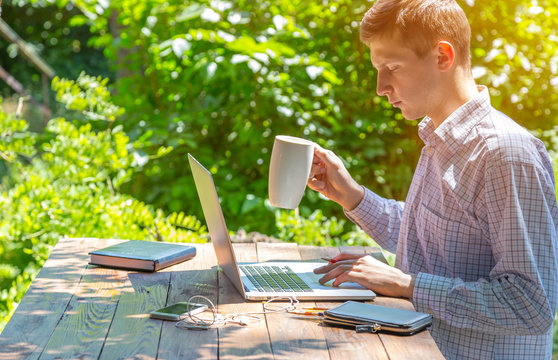 Freelancer working in park. Man sitting at rough country wooden desk working on computer drinking coffee business dress code electronic gadgets notepad around green forest sunbeams on background