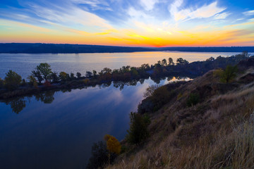 Gorgeous dawn on blue lake.
Luminous sunrise blooming over wild waterfront landscape with fall grass flower hill stone rocks and narrow island with colored trees in the middle of water surface