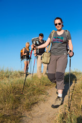 Family of hikers walks on clay path.
Three people two female one female heavy loaded with backpacks and trekking gear go along path throw wild grassy hill blue sky on background