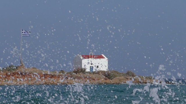 Zoomed view of a small separated from Europe mainland island in the sea with a little white church and Greek flag waving on a flagpole with waves splashing in the foreground. Conceptual clip