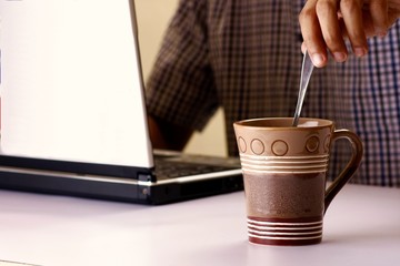 Fototapeta na wymiar Coffee in a mug being stirred by a man working on a laptop computer in the background