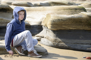 Young boy in blue jacket sitting on rocks gazing into the distance at the seaside. Taken in Whitley Bay in North East England
