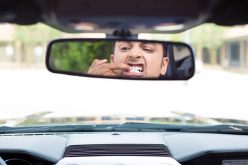 Closeup portrait, funny young man driver looking at rear view mirror to pick teeth with finger nails because of food stuck behind in between, isolated interior car windshield background
