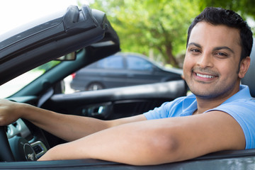 Closeup portrait, happy young smiling handsome man in blue polo shirt in his new black sports car, relaxing, looking at camera, isolated on outdoors background with vehicle and green trees.