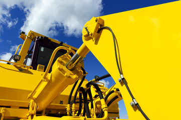 Bulldozer, huge yellow powerful construction machinery with big bucket, focused on hydraulic piston arm, blue sky and white clouds on background  - 86439458