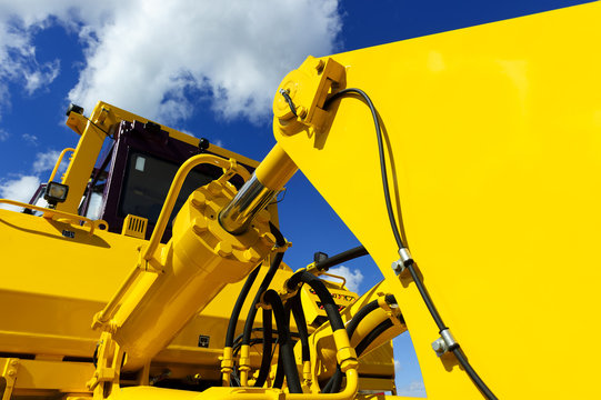 Bulldozer, Huge Yellow Powerful Construction Machinery With Big Bucket, Focused On Hydraulic Piston Arm, Blue Sky And White Clouds On Background 