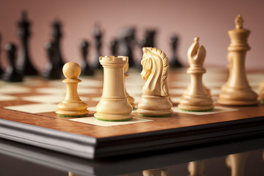 Traditional Staunton white chess pieces carved in natural boxwood (left pawn rook and knight in focus) standing on wooden superior chessboard and glossy table