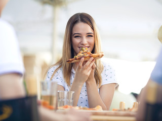 Cheerful  young woman enjoying slice of pizza while sitting with her friends in the pizzeria
