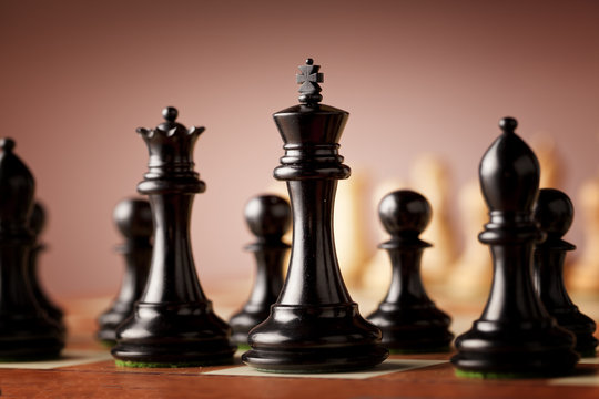 A line of luxurious black Staunton chess pieces carved in genuine ebony wood in focus standing traditional wooden chessboard facing the army of white pieces before the battle