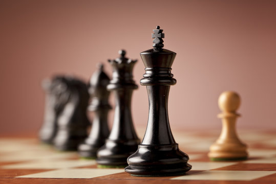 A line of luxurious chess pieces carved in genuine ebony wood facing a single white pawn made of natural boxwood standing on superior traditional wooden chessboard