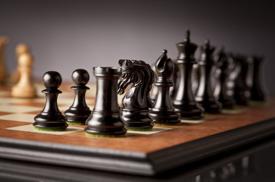 Left flank of the black army of luxurious chess pieces in focus on a wooden chessboard with dark background