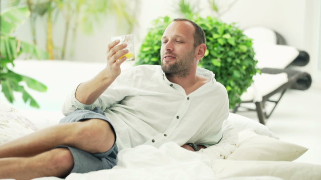 Happy, young man relaxing and drinking cocktail lying on bed on patio
