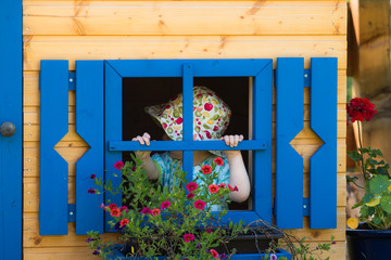 little girl with sunhat playing peekaboo in the kids playhouse with blue window and blooming flowers