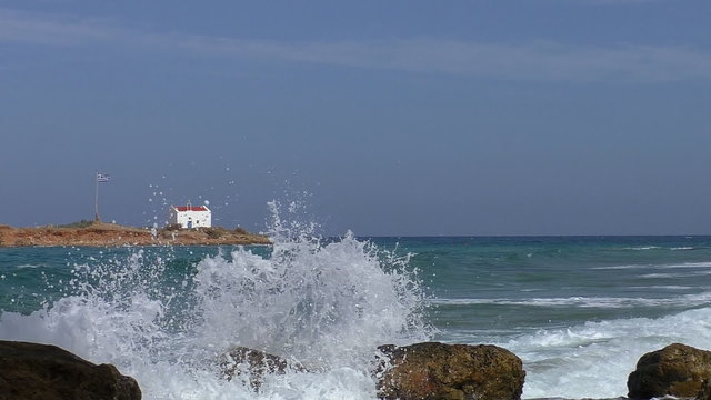 View of small island in the turquoise sea with white waves washing big stones in the foreground.Greek flag waving on flagpole and little white church on this separate piece of land of Greece. 