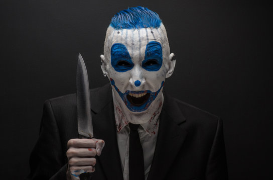Terrible clown and Halloween theme: Crazy blue clown in a black suit with a knife in his hand isolated on a dark background in the studio