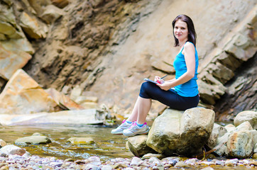 beautiful young girl sits on a rock in the river and draws from