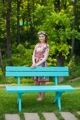 Beautiful young red-haired woman in a bright blue dress standing near a bench in the garden