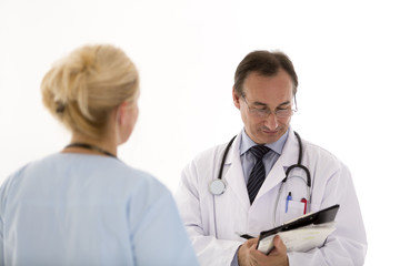 Male doctor with glasses writing something on his clipboard giving instructions to a blond female colleague, back turned. Indoors