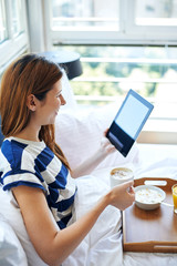 Young woman drinking coffee and reading digital tablet in bed