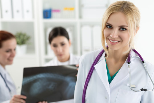 Portrait of smiling female medicine doctor holding blue document folder with two colleagues looking at x-ray picture at background. Healthcare and medicine concept.