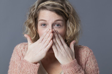 shy young blond woman with curly hair laughing behind her hands for fun and shyness