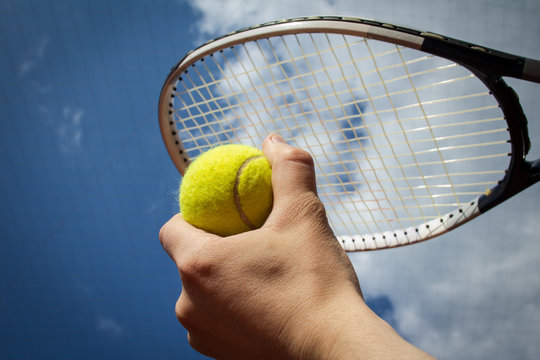 Hand Holding Tennis Ball And Racket Agaist Sky