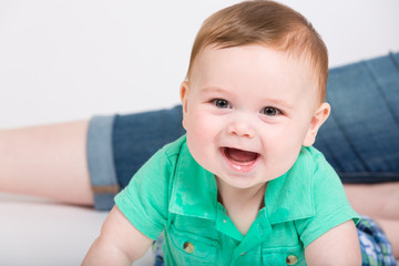 8 month year old baby lays on his stomach looking towards camera, while mom is sitting in background. dressed in a cute green polo shirt and blue plaid shorts.